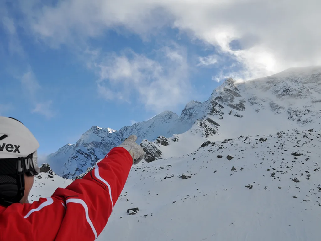 Person im Skihelm zeigt auf einen Gipfel unter Wolken.